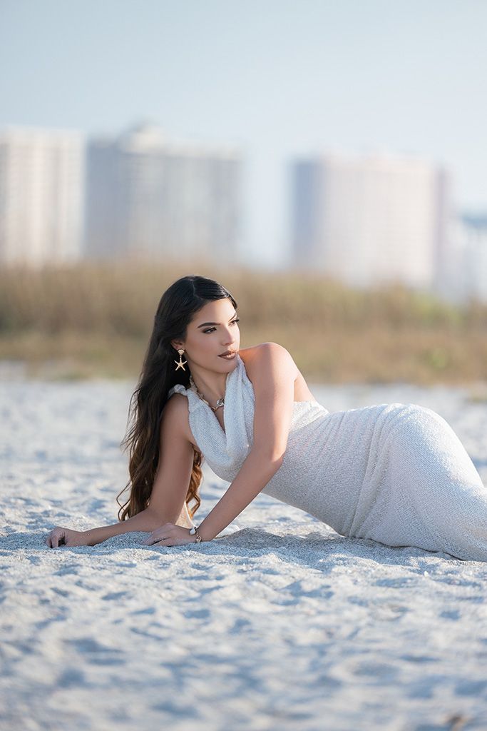 A woman in a white dress poses gracefully on white sand beach with coastal buildings in the background.