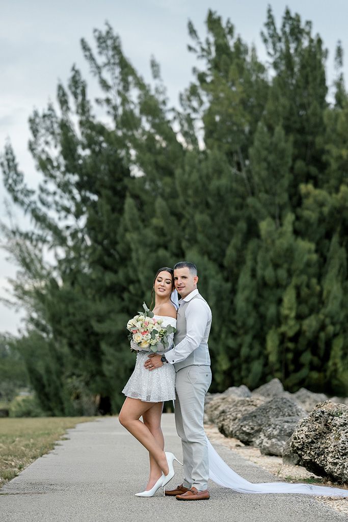 Bride and groom pose on a path surrounded by tall trees, bride holding a bouquet in a short white dress.