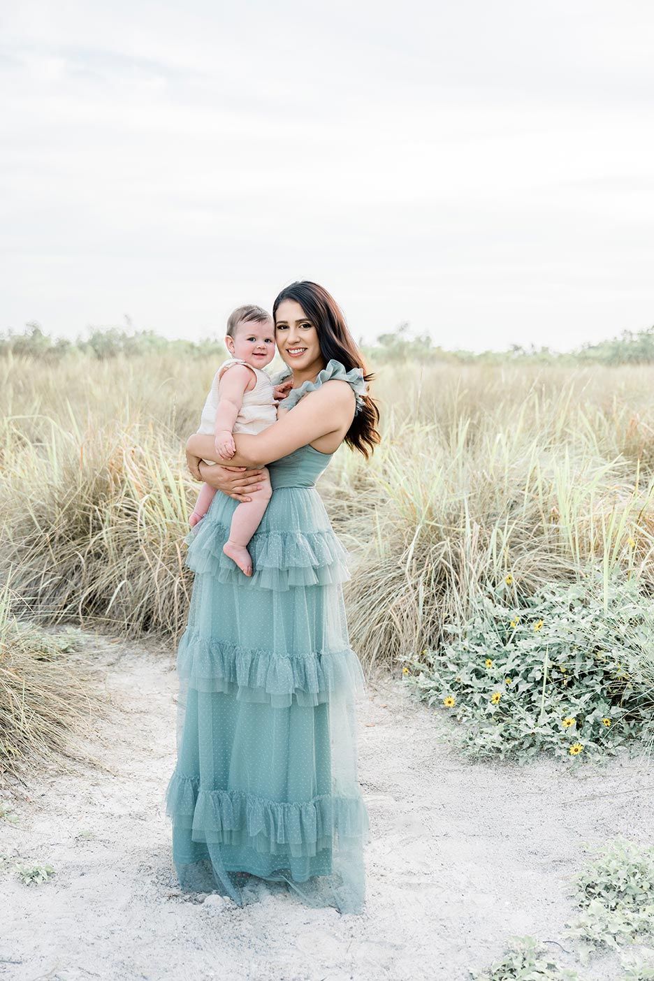A family in flowing blue dresses embraces on a sandy beach path surrounded by coastal vegetation in soft sunlight.