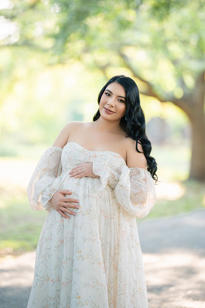 Pregnant woman in floral off-shoulder gown cradles baby bump outdoors under lush green trees in soft natural light.