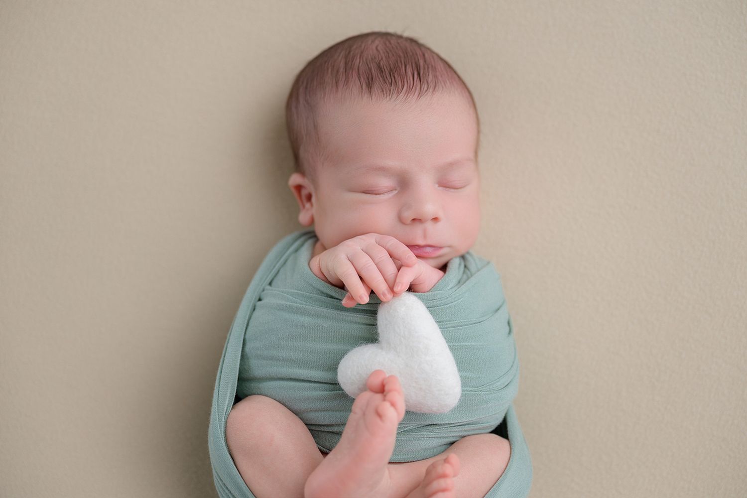 Newborn baby swaddled in teal wrap sleeping while holding a small white heart prop.