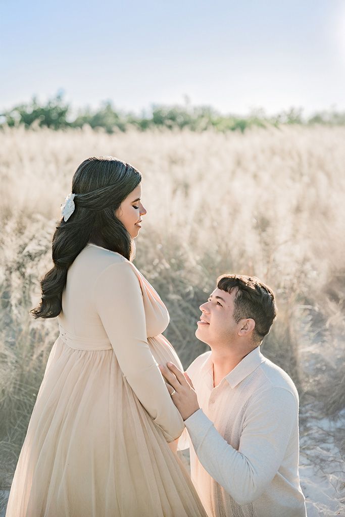 Couple sharing a tender maternity moment in a golden field, man kneeling touching pregnant womans belly in beige outfits.