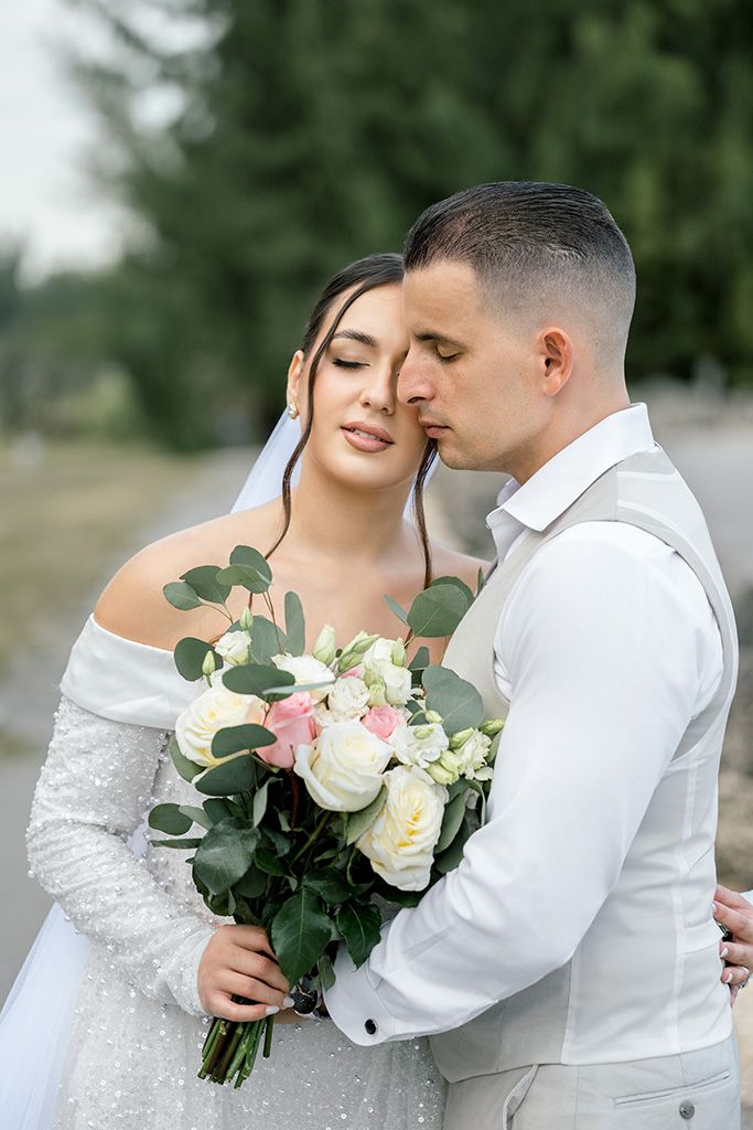 A bride in a white beaded gown holds a rose bouquet while the groom in a white suit tenderly kisses her cheek outdoors.