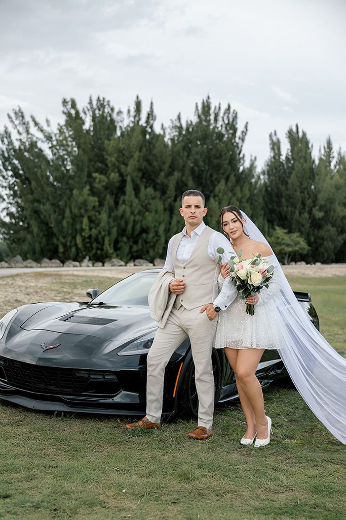 Bride and groom posing in front of a black Corvette sports car on a grassy field with trees in the background.