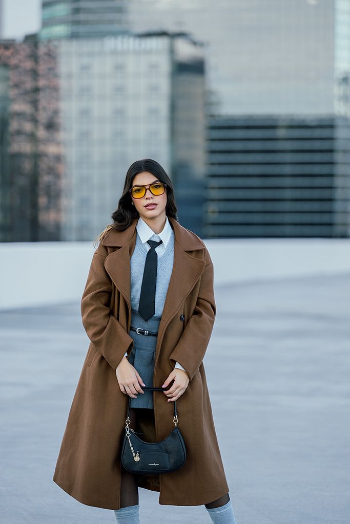 Woman in brown coat, gray suit, black tie, yellow sunglasses, holding black handbag, standing outdoors with city buildings.