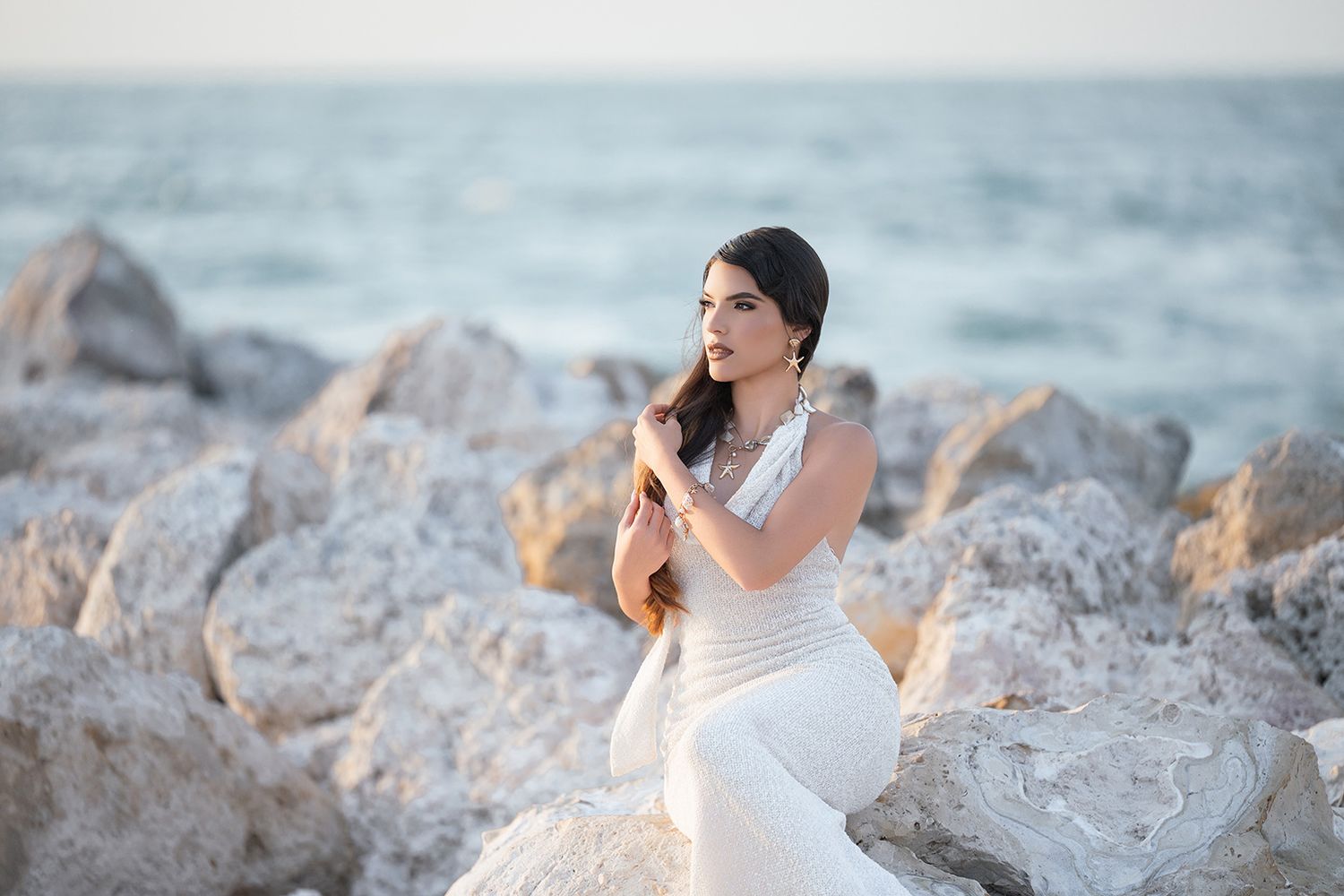 Woman in white dress sitting on rocks by the ocean at sunset, hands clasped near chest.
