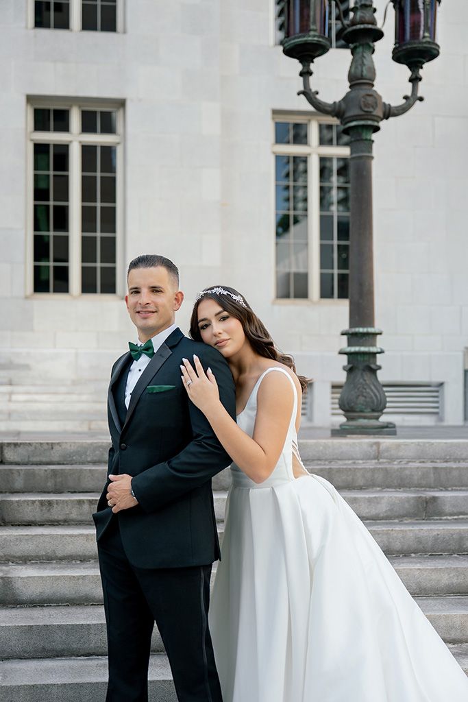 Bride in white ballgown leans on groom in black tuxedo on stone steps outside elegant building.