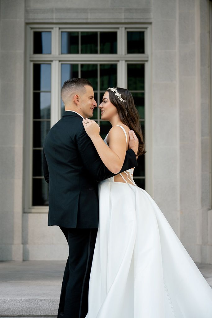 Bride and groom embracing outside elegant stone building, bride in backless white ball gown.
