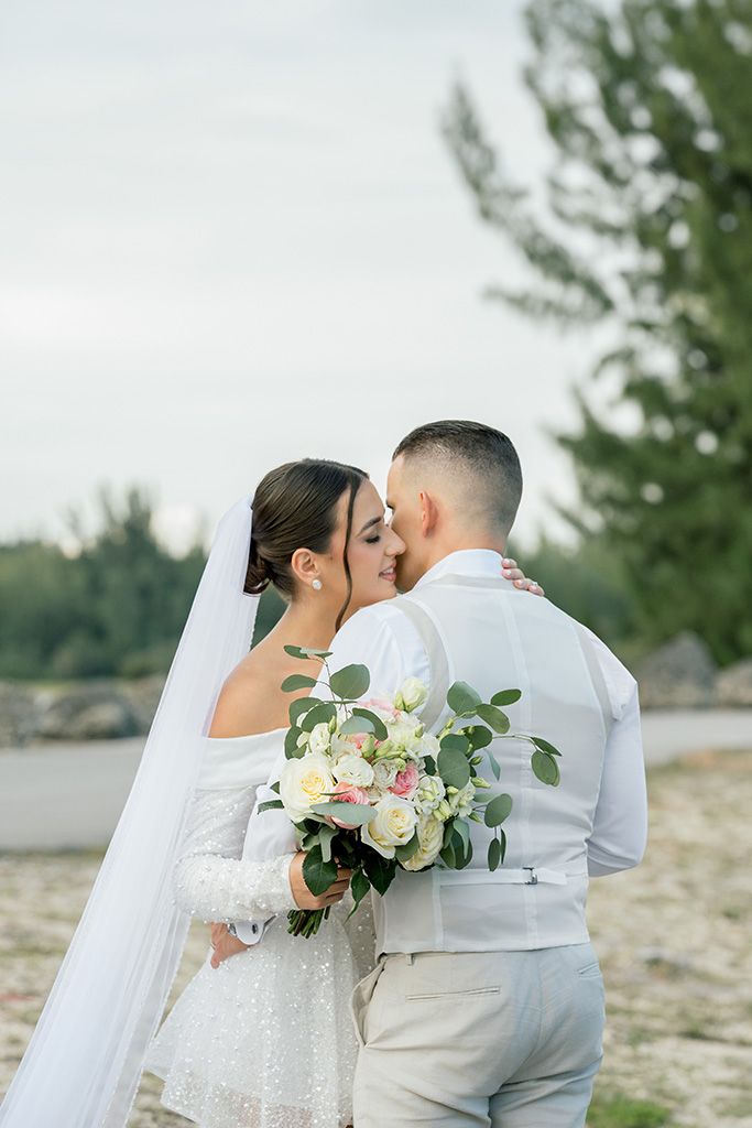 Bride and groom sharing a tender forehead touch, bride holding white rose bouquet with veil, outdoor beach wedding.