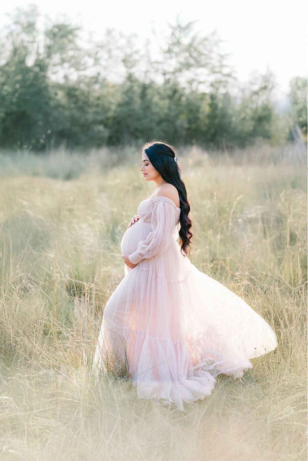 Ethereal maternity photo in a dreamy field shows an expectant mother in a flowing pale pink off-shoulder gown at sunset.