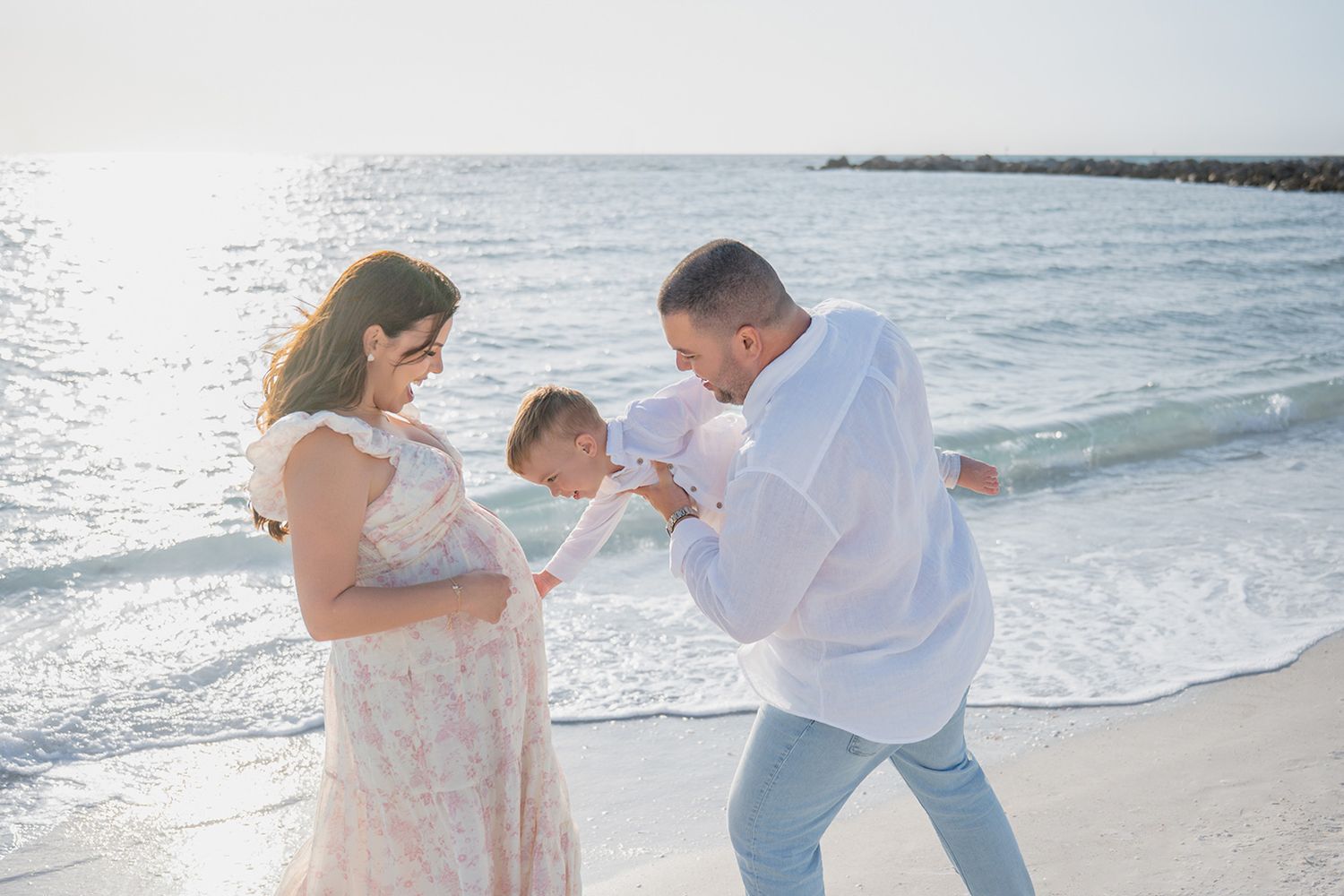 Family of three enjoying a sunny beach day, with parents playfully engaging with their toddler by the ocean waves.