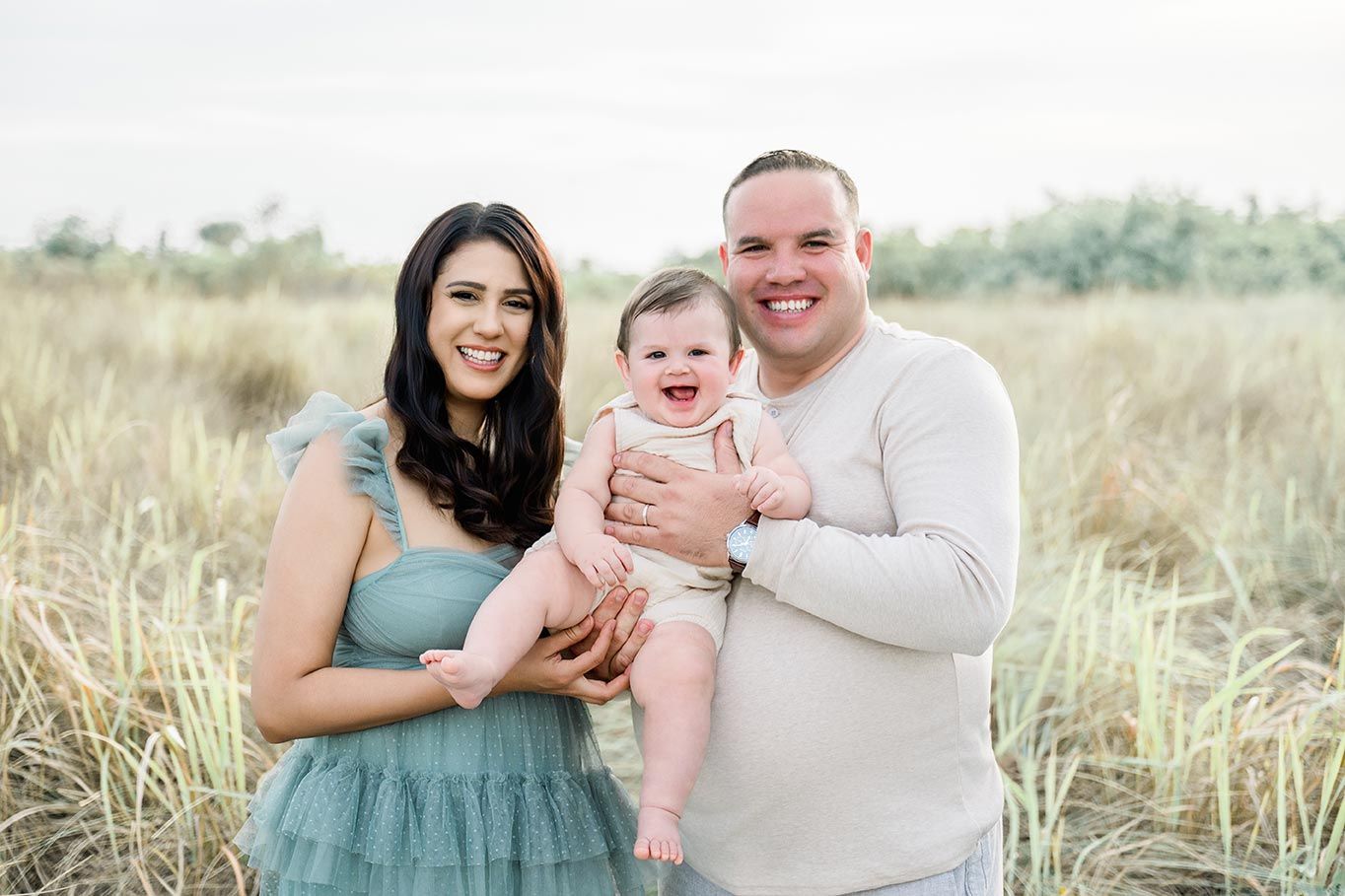 Happy family of three posing in golden grass field, parents holding smiling baby in pink outfit, outdoor portrait.