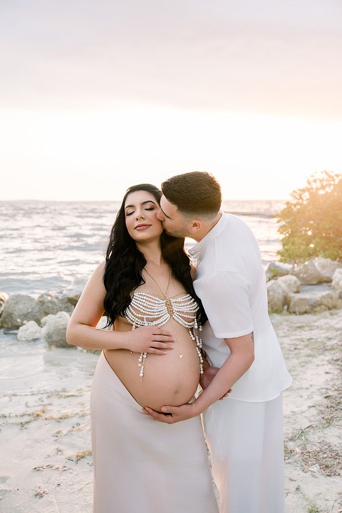 Couple shares tender maternity beach moment at sunset, man kissing pregnant woman in white beaded dress by rocky shore.