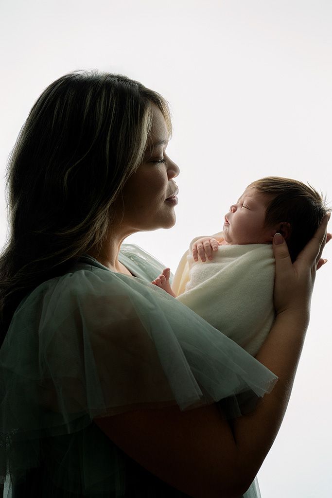 A mother in a teal gown tenderly holds her sleeping newborn baby against a soft white background.