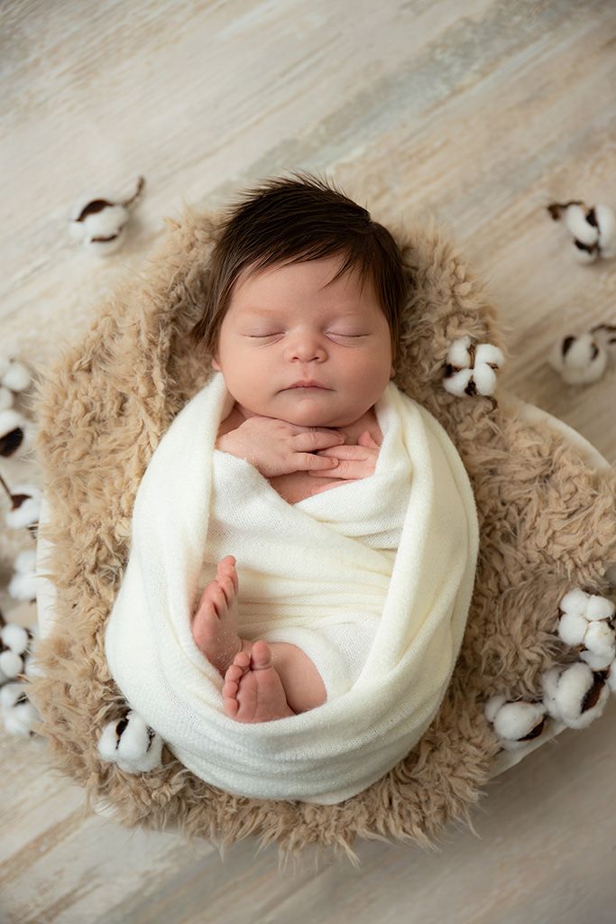 Sleeping newborn baby swaddled in white wrap resting in a fuzzy bowl surrounded by cotton flowers.