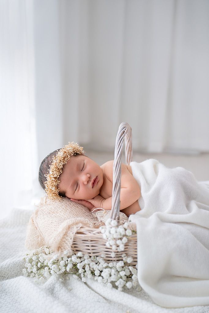 Peaceful newborn baby sleeping in white wicker basket with floral crown and white blanket and flowers.