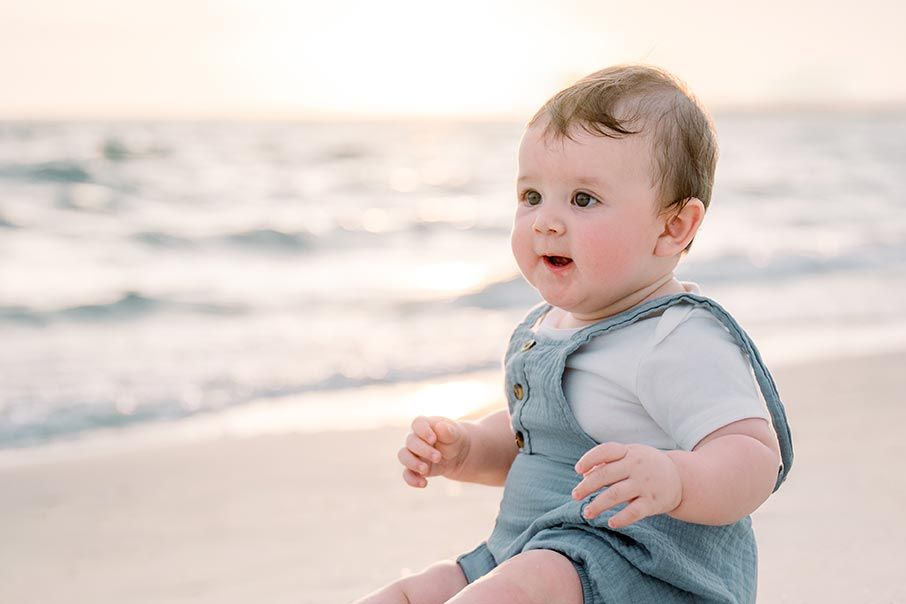 A small baby in blue sits on a sandy beach during sunset with a serene coastal view.