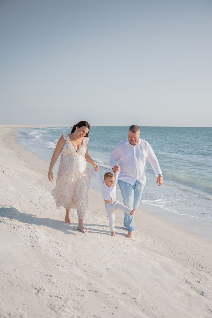 A happy family walks along a pristine white sand beach, parents holding their toddlers hands by the sea.