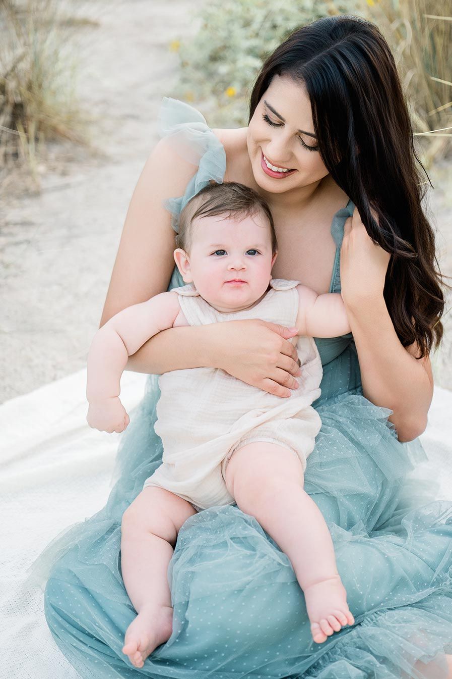 A smiling brunette woman in a blue tulle dress holds her happy baby outdoors for a sweet mother and child portrait.