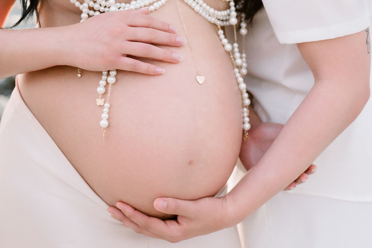 Pregnant woman in white dress adorned with pearl necklace draped over baby bump, partner embracing from behind.