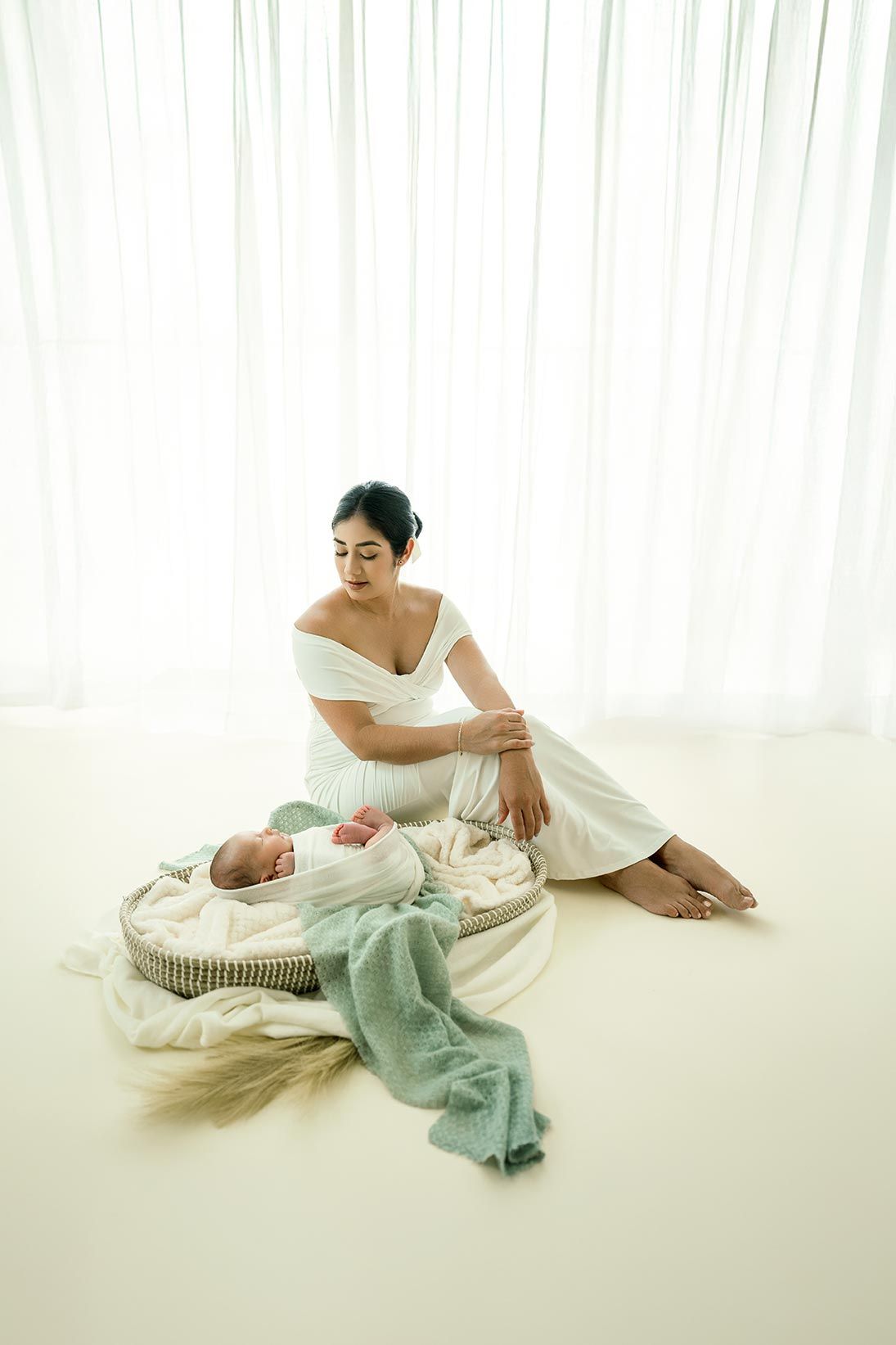 Mother in white dress sitting beside sleeping newborn baby in woven basket with soft blankets by sheer curtains.