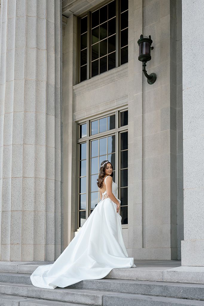 Bride in flowing white gown poses gracefully on stone steps between tall columns of an elegant building.