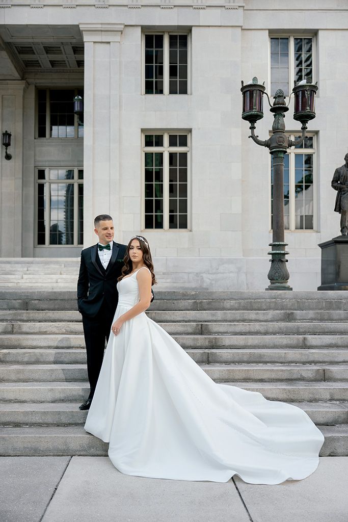 Bride in white ball gown and groom in black tuxedo pose on steps of elegant stone building.