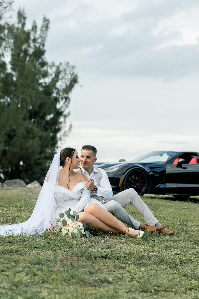Newlywed couple sitting on grass by a black sports car, bride in white gown with veil holding bouquet.