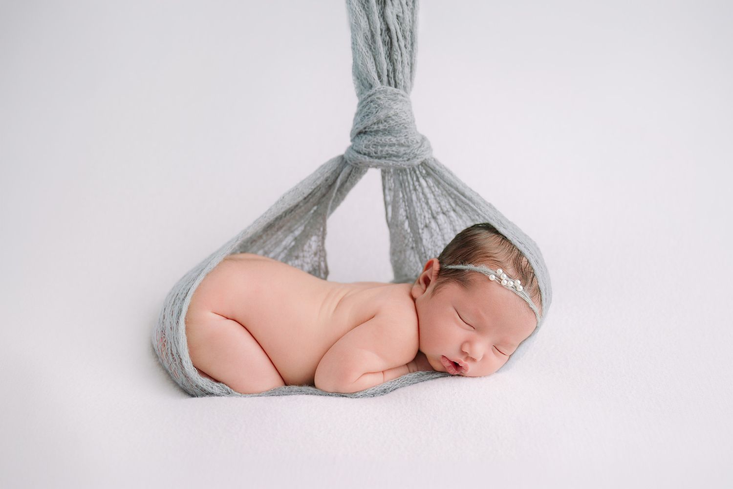 Sleeping newborn baby girl cradled in a soft gray wrap suspended against a white background.