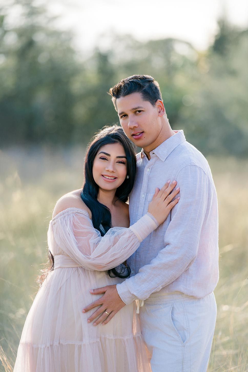 A couple embraces lovingly in soft natural lighting against a dreamy outdoor backdrop in pastel colors.