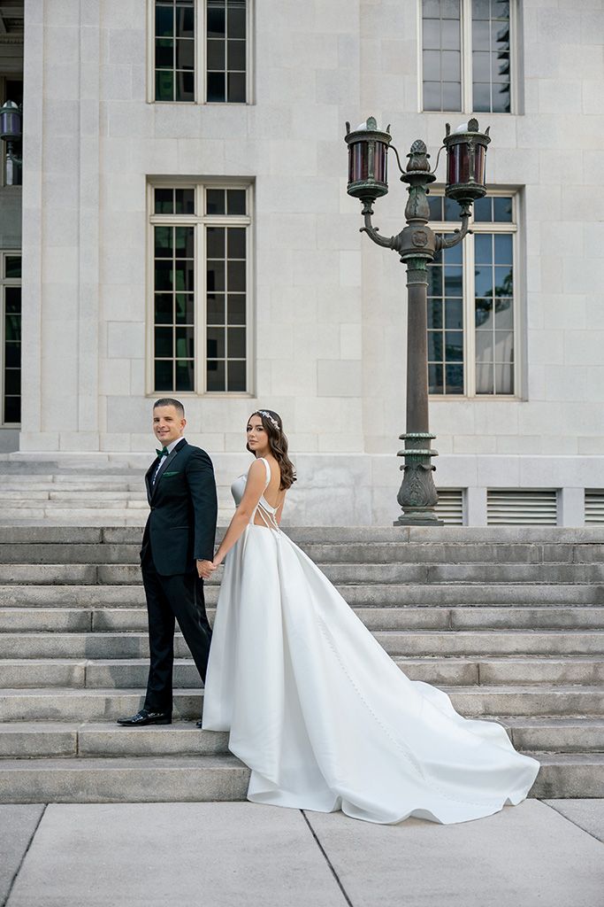 Bride and groom holding hands on stone steps outside a grand building, bride wearing a flowing white gown with long train.