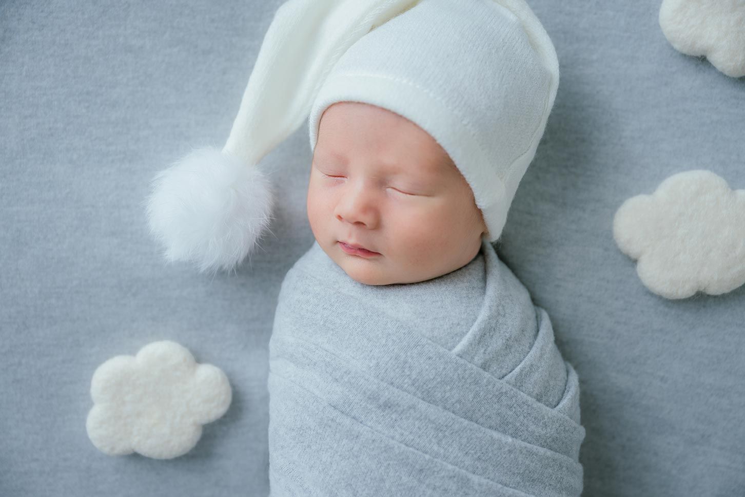 Sleeping newborn baby swaddled in gray wrap wearing white pompom hat surrounded by cloud decorations.