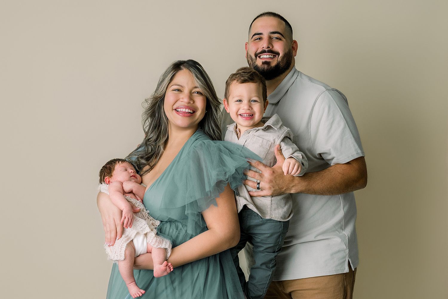 Family portrait of parents holding newborn baby and toddler son, smiling against neutral backdrop.