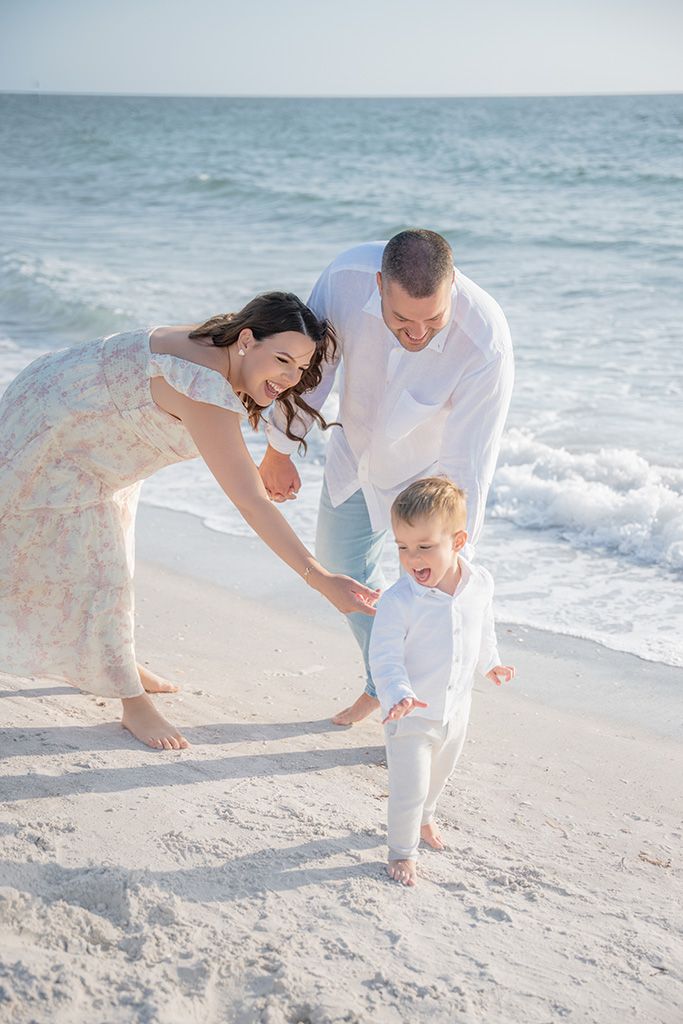 Happy parents in white outfits helping their laughing toddler walk along a bright sandy beach shoreline.