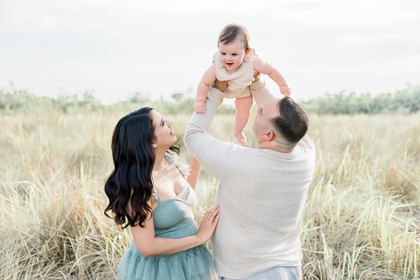 Parents lifting happy baby in sunny field, family portrait photography outdoors.