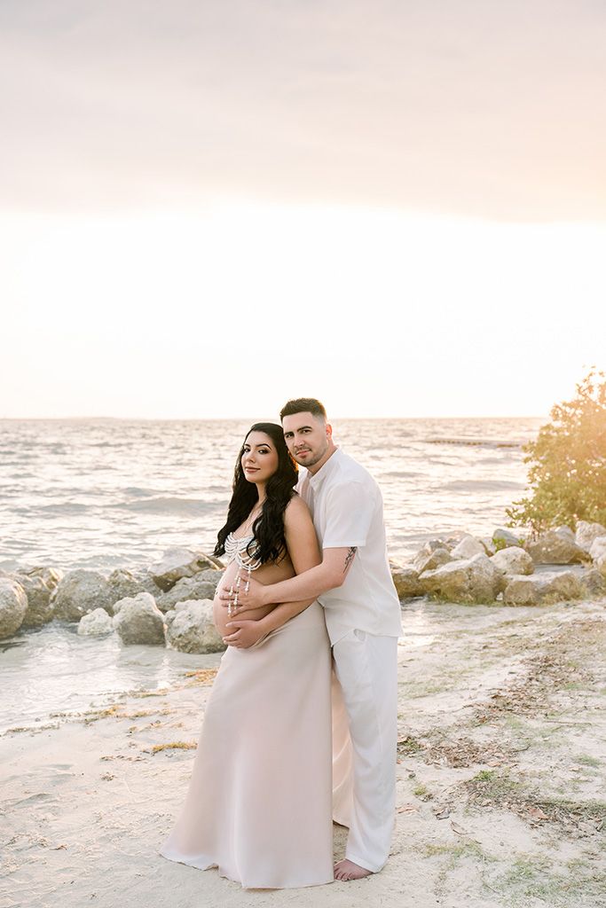 Couple embracing on a sandy beach at sunset, woman in blush gown cradling baby bump, man in white outfit behind her.