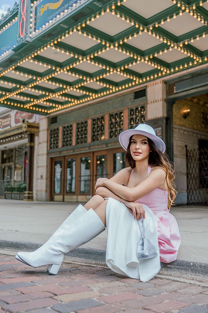 Woman in pink top, white skirt, knee-high boots and lavender hat sits outside a lit theater.