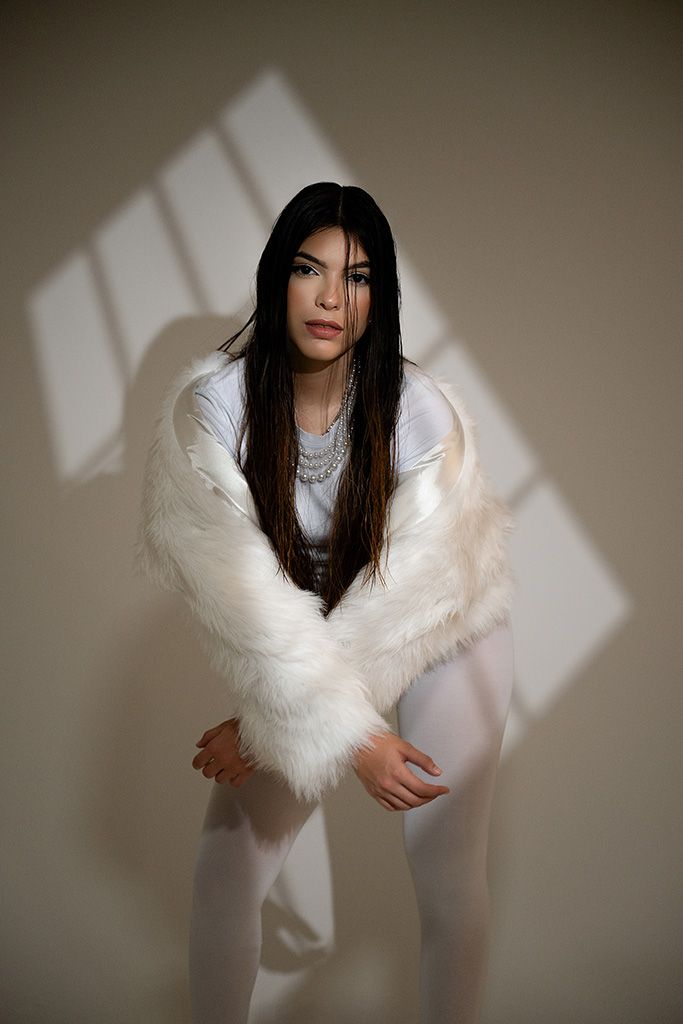 Woman in white faux fur jacket and pants poses elegantly with silver necklace in a softly lit studio.