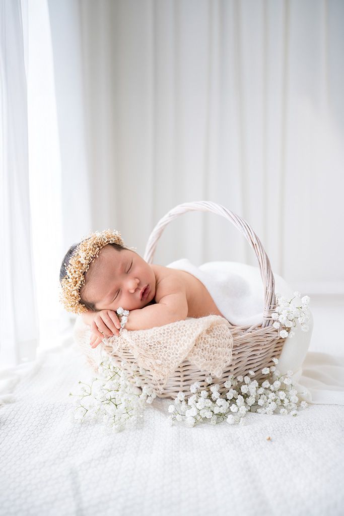Peaceful newborn baby sleeping in white wicker basket with floral crown and white baby's breath flowers.