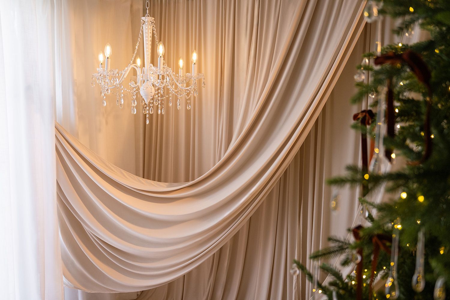 Elegant room with draped cream curtains, crystal chandelier, and decorated Christmas tree with warm glowing lights.