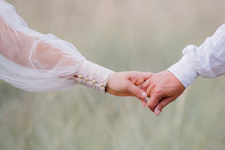 Two hands reaching out and holding each other with soft focus and ethereal white clothing in dreamy outdoor setting.