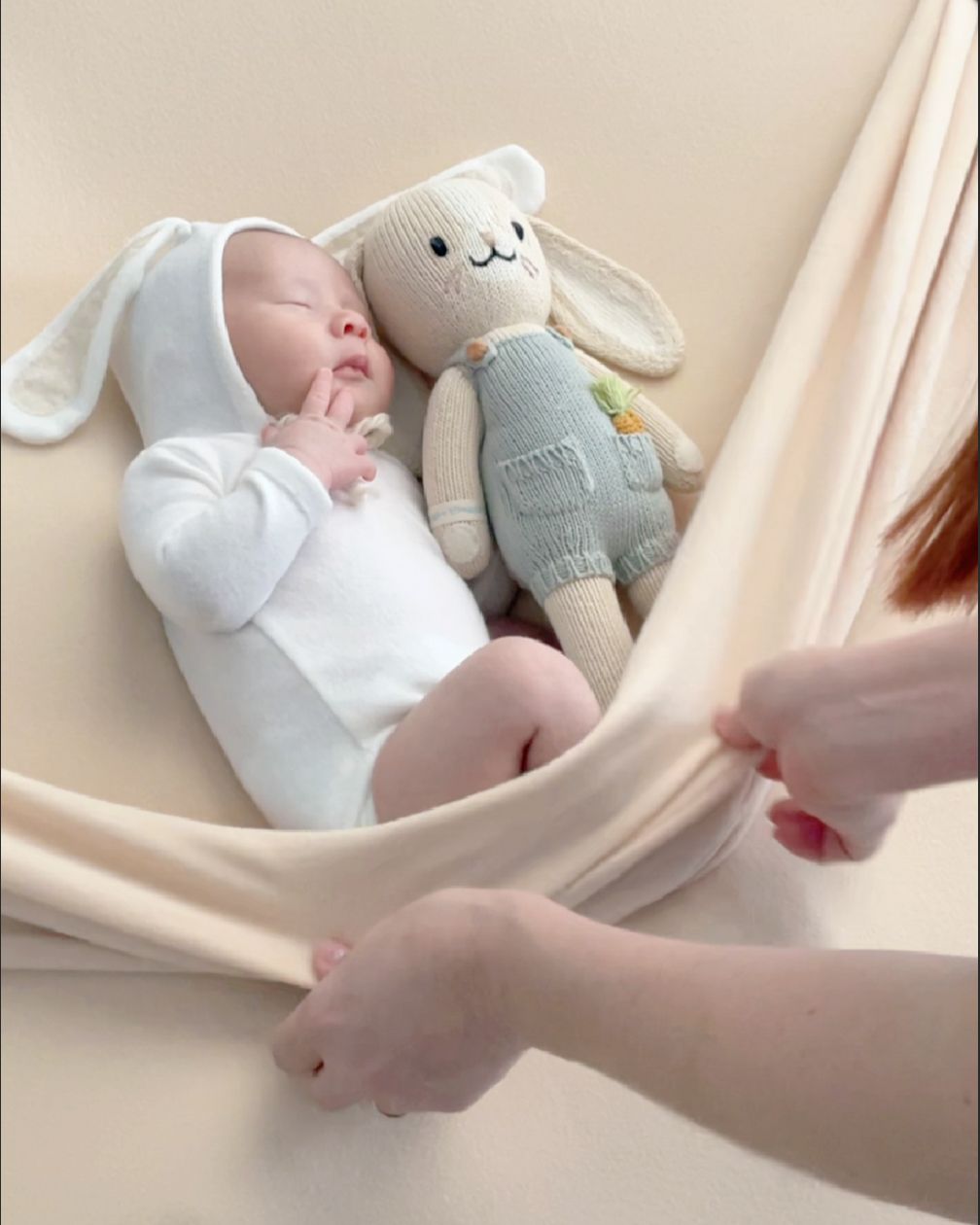 A small baby lies on a cream-colored fabric hammock next to a soft gray stuffed bunny toy wearing blue overalls.