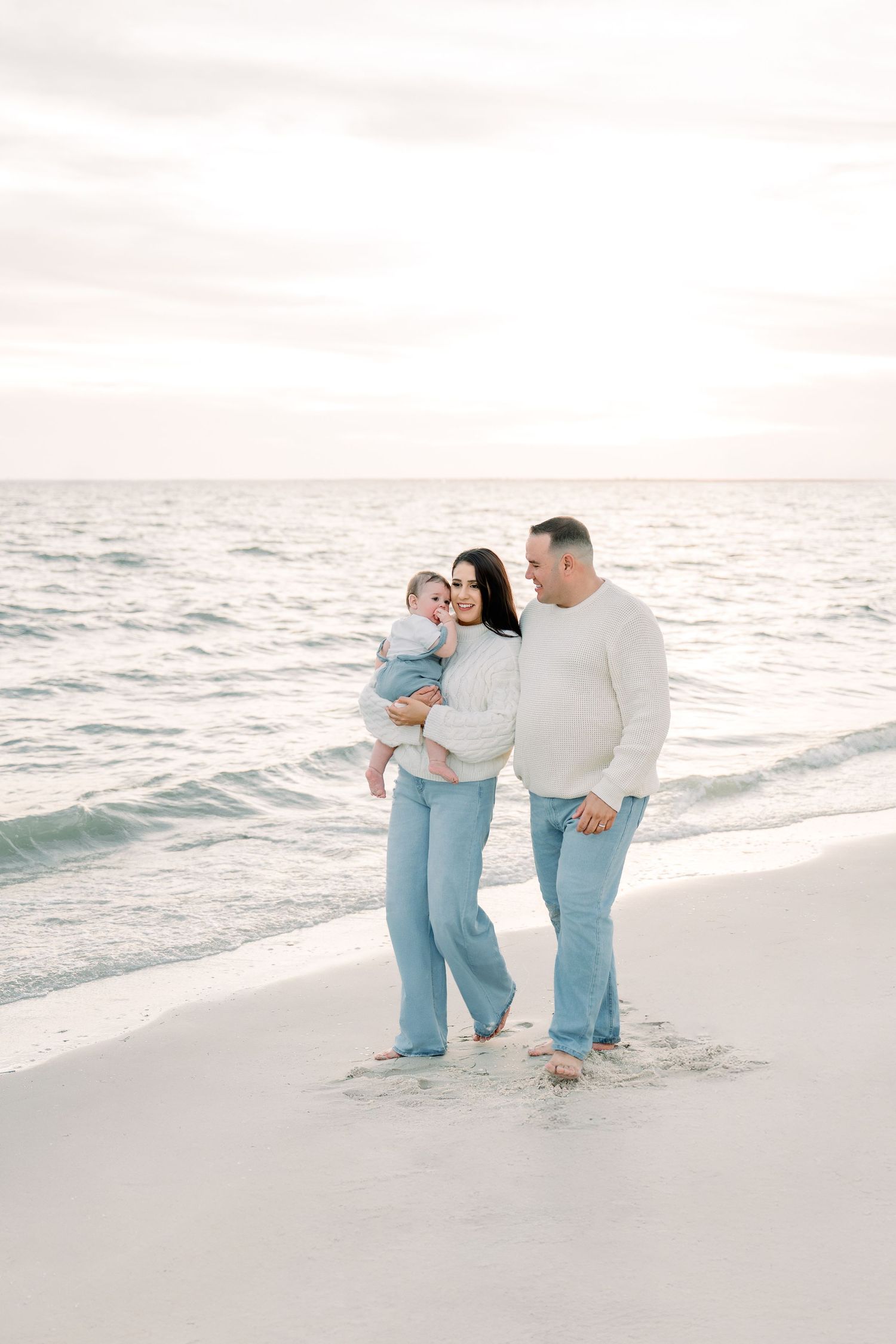 A family in matching light blue and white outfits walks along a serene beach at sunset, creating a dreamy coastal scene.