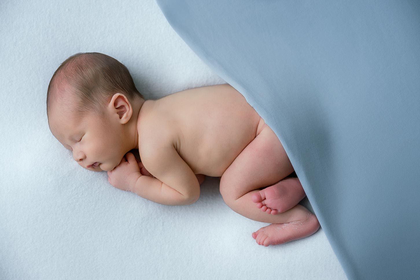 Newborn baby sleeping peacefully curled up on a soft blue blanket in a studio photography setting.