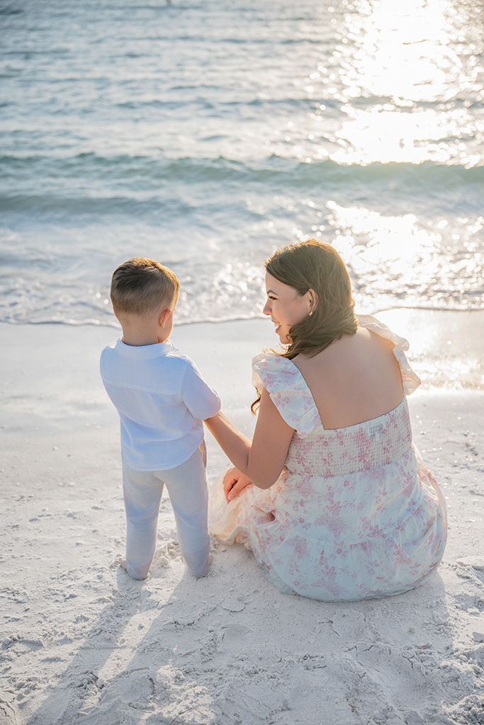 A mother and young son sit together on a sandy beach at sunset, sharing a tender moment by the ocean waves.