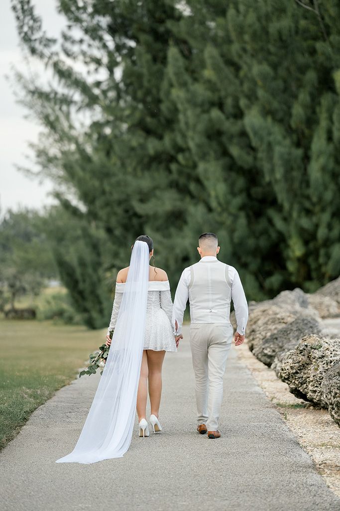 Bride and groom walk hand in hand along a scenic path lined with tall green trees on their wedding day.