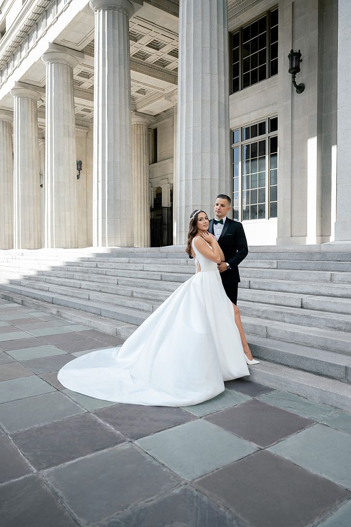 Bride in white ballgown and groom in tuxedo pose on grand stone steps with tall columns.