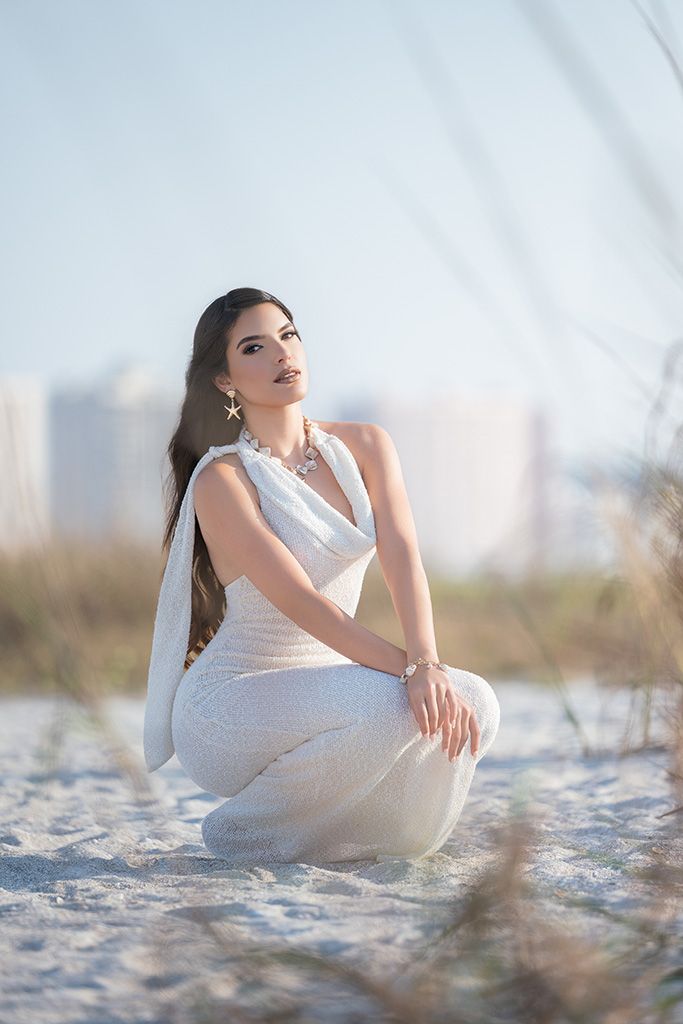 Beautiful brunette woman in a white sequin gown sitting gracefully on white sand dunes with a coastal skyline.