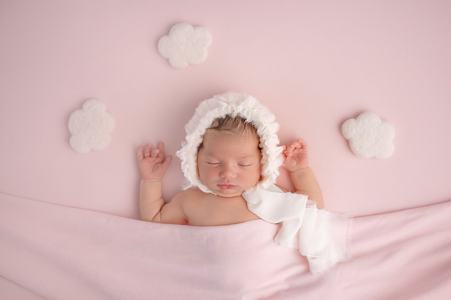 Newborn baby sleeping peacefully on pink blanket wearing white bonnet, surrounded by fluffy cloud decorations.