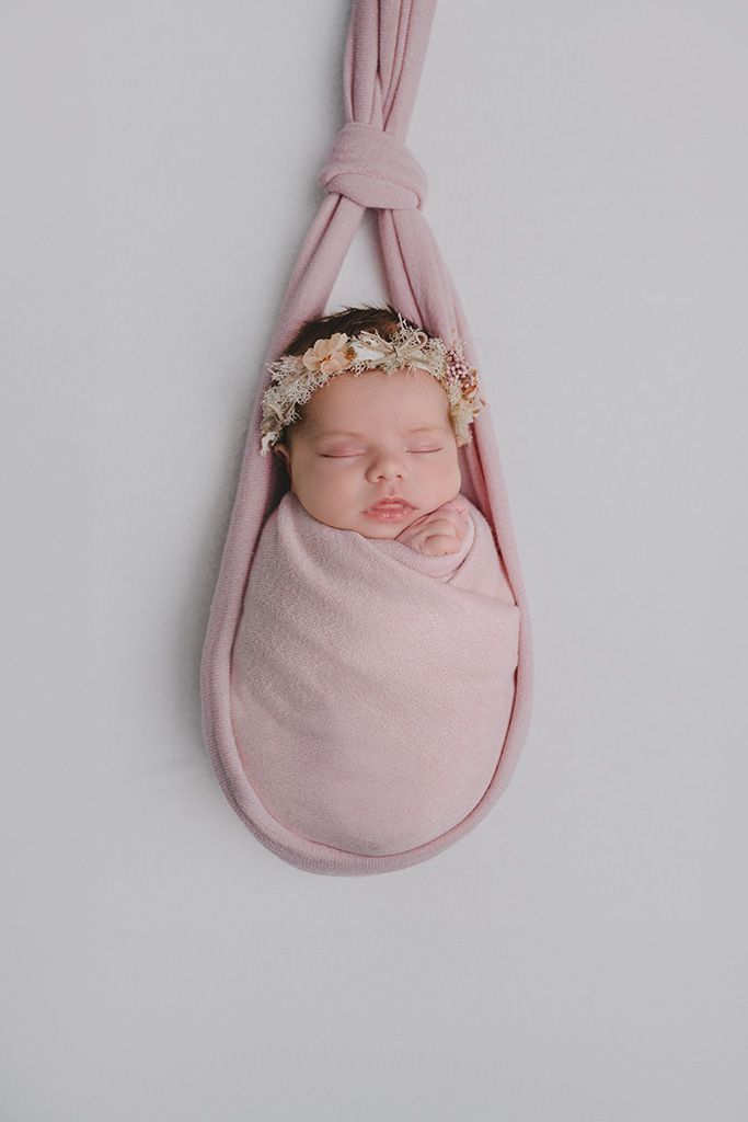 Sleeping newborn baby girl swaddled in pink wrap with floral crown, posed in hanging fabric sling.
